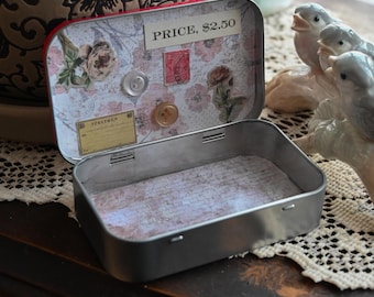 May include: An open, rectangular metal tin with a red rim, decorated with floral and vintage paper designs. The interior features a collage of stamps, buttons, and a price tag. Decorative birds and a vase are in the background.
