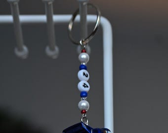 May include: A blue toy car with white and red accents, hanging from a white metal stand. The car has the number 24 on the side and the words "Audi Sport" on the back. The car is attached to a keychain with white and blue beads.