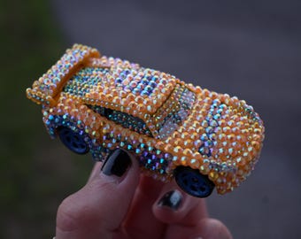 May include: A toy car covered in iridescent orange and blue rhinestones. The vehicle features a rear spoiler, a roof section, and dark blue wheels. The surface is entirely encrusted with small, reflective crystals that catch the light.