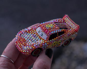 May include: A miniature toy race car completely covered in iridescent, multi-colored rhinestones that reflect light in shades of red, orange, and yellow. The car features a rear spoiler, black wheels with white lettering, and a windshield with printed eyes.