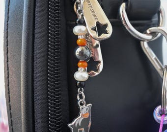 May include: A silver-toned metal zipper pull charm attached to a black bag zipper. The charm features a vertical arrangement of small black, white, and orange beads, ending in a silver-toned metal pendant shaped like a cat with orange accents.