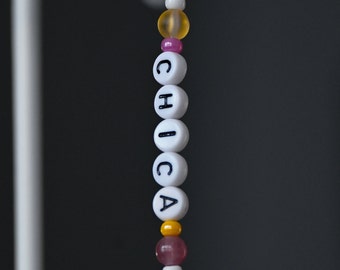 May include: A beaded keychain spelling "CHICA" in white letter beads. The keychain features a silver ring, with yellow, pink, and clear beads. The background is a dark gray.