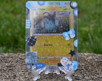 May include: A Shinx Pokémon trading card, featuring a blue and black creature, is displayed in a clear protective case. The card is decorated with blue and black embellishments, including a bow and star, and is propped up on a clear stand.