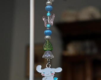 May include: A beaded keychain with a silver ring. The keychain features blue and pearl beads, a pink butterfly charm, a green leaf charm, a purple flower charm, and a light blue cartoon character on a skateboard.