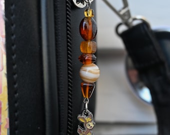 May include: A beaded charm with a deer pendant hangs from a black bag. The charm features a series of amber and white beads, with a small deer charm at the bottom. The bag has a silver zipper and a black strap.