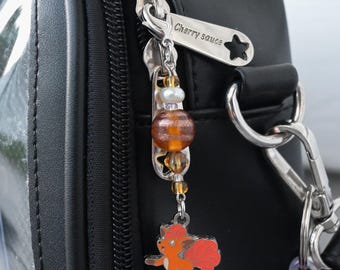 May include: A black bag with a silver zipper pull engraved with the text Cherry sauce and a star cutout. Attached to the zipper is a charm featuring a small orange fox-like creature, a white pearl bead, and two amber-colored striped glass beads.