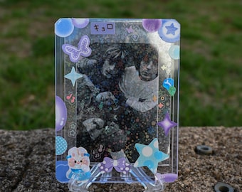May include: A clear, rectangular photo frame with a black and white image of two people. The frame is decorated with purple, blue, and white stickers, including stars, butterflies, and a cartoon character. The frame is supported by a clear plastic stand.