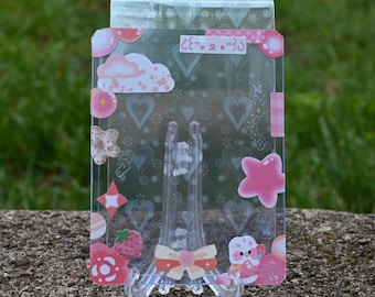 May include: A clear, rectangular plastic card holder decorated with pink and white stickers of clouds, hearts, stars, and a strawberry. The holder is displayed on a clear plastic stand, set against a green background.
