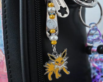 May include: A zipper pull charm attached to a black bag, featuring a yellow and white beaded strand with a metal Jolteon character pendant. The silver-toned zipper pull has a star-shaped cutout and the text "arty sauce" engraved on its surface.