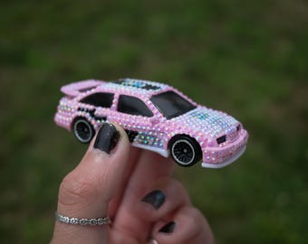 May include: A miniature toy car covered in small, light pink, iridescent rhinestones. The vehicle features black wheels, a rear spoiler, and dark-tinted windows. The body of the car is completely encrusted with sparkling gems, creating a textured, reflective surface across the entire exterior of the model car.