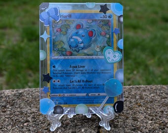 May include: A Pokémon trading card featuring Marill, a blue creature, with the text "Aqua Liner" and "Let's All Rollout." The card is displayed in a clear protective case decorated with blue accents and star shapes. The card is on a clear stand.