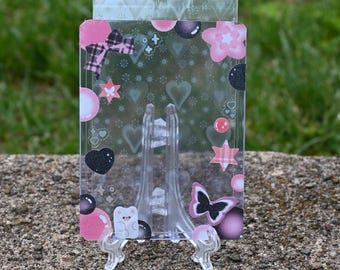 May include: A clear acrylic display stand with a pink and black decorative design. The design includes hearts, stars, bows, and a butterfly. The stand is supported by a clear plastic easel.