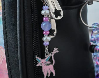 May include: A beaded zipper pull charm attached to a black bag zipper. The charm features a pink and teal Espeon character pendant hanging below a sequence of small white pearls, translucent purple beads, and metallic pink spacer beads.