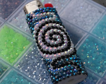 May include: A cylindrical lighter covered in small, reflective rhinestones arranged in a spiral pattern. The design features alternating rows of black, iridescent white, and deep blue crystals, with a metallic top and a small red ignition button.
