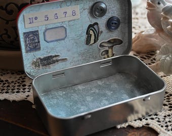 May include: A rectangular metal tin with an open lid featuring a light blue map background, a label with the text N° 5 6 7 8, a postage stamp, a butterfly sticker, a mushroom sticker, and two dark buttons attached to the interior lid surface.