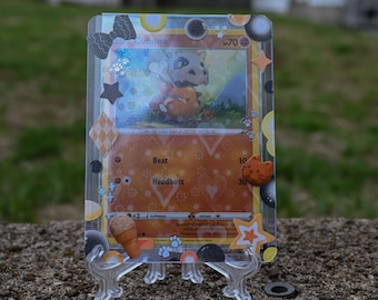 May include: A Cubone Pokémon trading card, displayed in a clear protective case. The card features an illustration of Cubone, with attack details. The case is decorated with stickers, including stars and a bow. The card is propped up on a clear plastic stand.