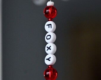 May include: A white beaded keychain with the word "FOXY" spelled out in black letters. The keychain is attached to a silver key ring and has red beads on either end.