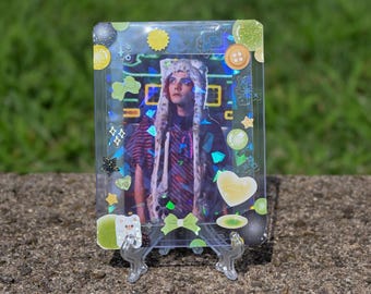 May include: A rectangular plastic card holder containing a photograph of a person wearing a white faux fur hat with long ear flaps. The holder is decorated with various stickers including green bows, yellow hearts, and buttons, with a holographic finish.