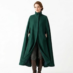 May include: A floor-length forest green cape coat featuring a high collar, a concealed front button closure, and arm slits. The garment is worn over dark gray trousers and brown leather knee-high boots, creating a structured silhouette.