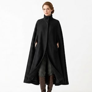 May include: A floor-length black wool cape featuring a high stand-up collar, a concealed front button closure, and side arm slits. The garment has a structured silhouette with clean lines, extending down to the ankles, worn over dark trousers and boots.