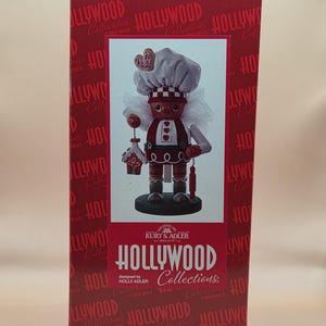 May include: A decorative nutcracker figurine from the Hollywood Collections, designed by Holly Adler. The figurine is dressed as a chef, holding a gingerbread house and a candy cane. The box is red with the words "Hollywood Collections".