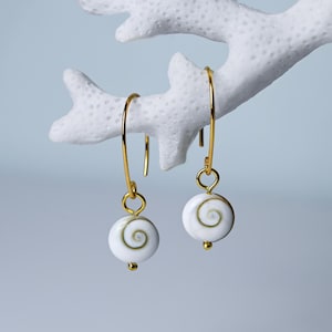 Könnte beinhalten: Goldfarbene Ohrringe mit weißen Anhängern im Spiraldesign. Die Ohrringe hängen an dünnen, gebogenen Golddrähten. Die Anhänger haben eine kleine goldene Perle am unteren Ende. Präsentiert vor einer weißen, korallenartigen Struktur.