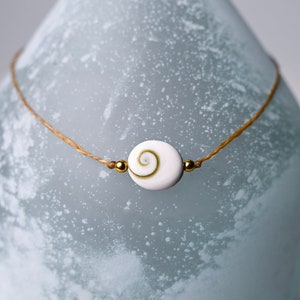 May include: A simple necklace with a white shell pendant and gold beads on a brown cord. The necklace is displayed on a frosted glass vase.