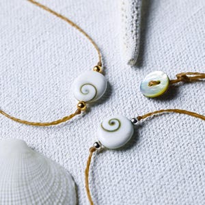 May include: Two bracelets with white shell beads and brown cords. The beads have a spiral pattern inside. One bracelet has a gold accent and the other has a silver accent.