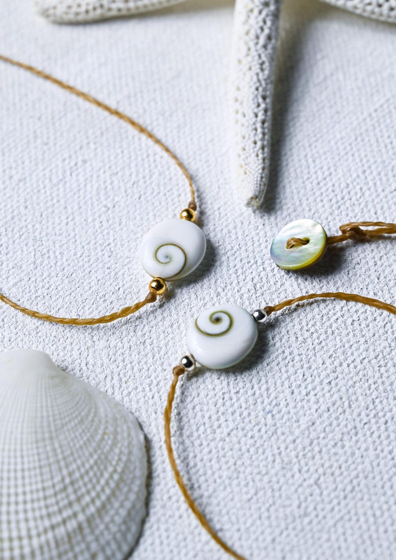 May include: Two bracelets with white shell beads and brown cords. The beads have a spiral pattern inside. One bracelet has a gold accent and the other has a silver accent.