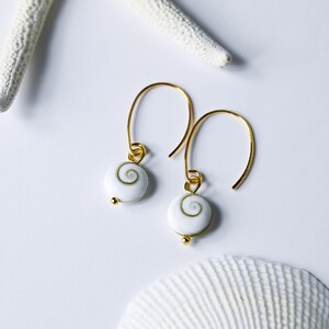 Op de afbeelding: Gouden oorhangers met witte kralen met een spiraalpatroon. De oorbellen worden gepresenteerd op een witte ondergrond, vergezeld van een zeester en zeeschelp, wat een maritiem thema oproept.