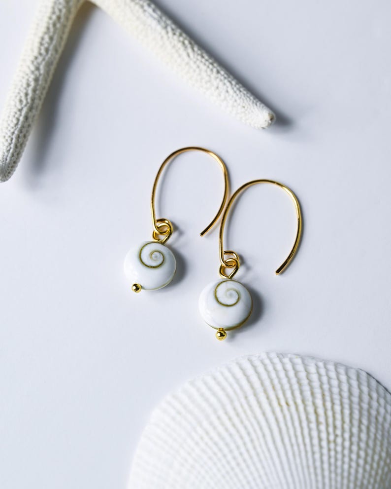 Op de afbeelding: Gouden oorhangers met witte kralen met een spiraalpatroon. De oorbellen worden gepresenteerd op een witte ondergrond, vergezeld van een zeester en zeeschelp, wat een maritiem thema oproept.