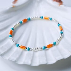 May include: A delicate bracelet featuring white pearl beads, turquoise blue beads, and orange rectangular beads. The bracelet is displayed on a white seashell.