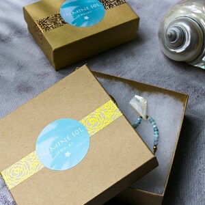 Op de afbeelding: Twee open bruine sieradendoosjes met het "Jasmine Sol Jewelry" logo op het deksel. E&eacute;n doosje bevat een kralenarmband met een witte schelpenhanger. Op de achtergrond een zeeschelp.