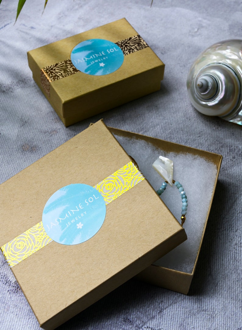Op de afbeelding: Twee open bruine sieradendoosjes met het "Jasmine Sol Jewelry" logo op het deksel. E&eacute;n doosje bevat een kralenarmband met een witte schelpenhanger. Op de achtergrond een zeeschelp.