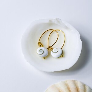 Op de afbeelding: Gouden oorringen met witte schelpenbedels met een spiraalpatroon. De oorbellen liggen in een witte schelp tegen een witte achtergrond. Kleine gouden details.