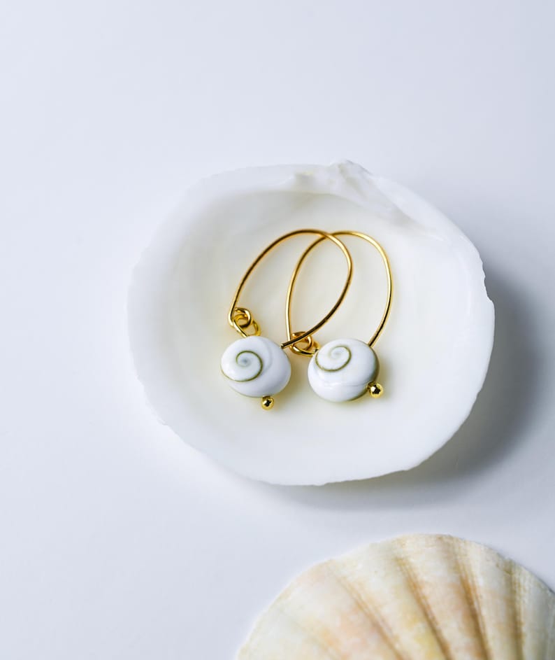 Op de afbeelding: Gouden oorringen met witte schelpenbedels met een spiraalpatroon. De oorbellen liggen in een witte schelp tegen een witte achtergrond. Kleine gouden details.