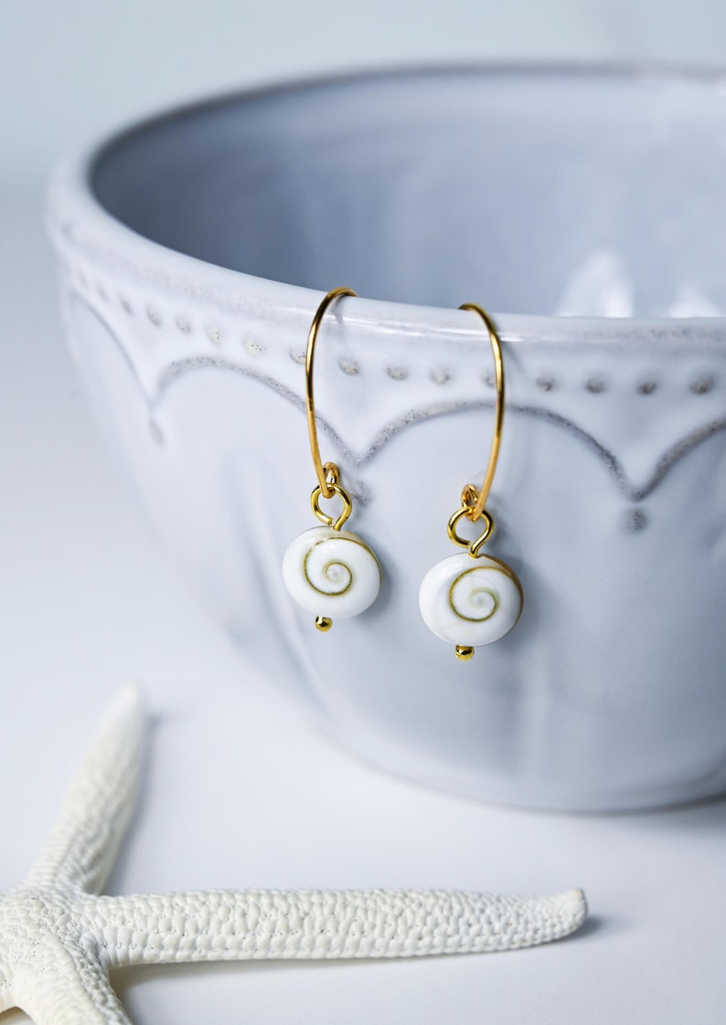 Op de afbeelding: Gouden oorringen met witte, spiraalvormige hangers. De oorbellen hebben een kleine gouden kraal aan de onderkant van de hanger. De oorbellen worden getoond in de buurt van een witte kom en een zeester.