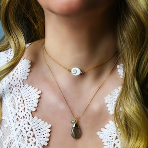 May include: Two gold necklaces with pendants. The top necklace has a white oval pendant with a green swirl design. The bottom necklace has a teardrop-shaped pendant with a gray stone and a small green bead.