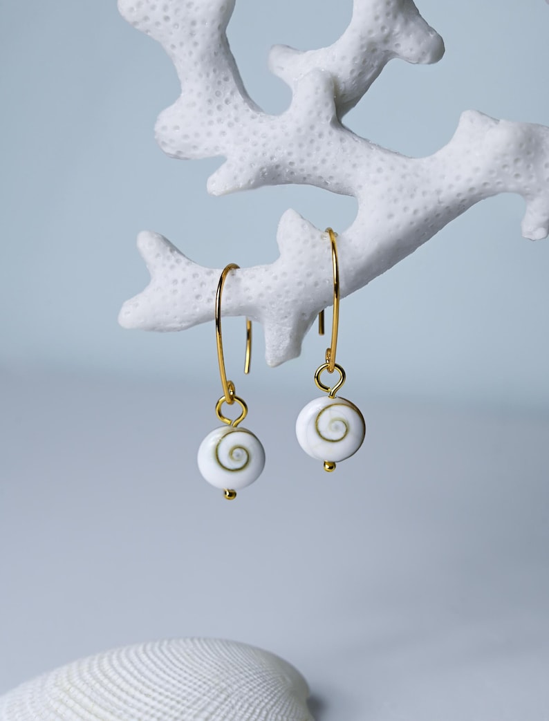 Op de afbeelding: Goudkleurige oorbellen met witte, spiraalvormige hangers. De oorbellen hangen aan dunne, gebogen gouden draden. De hangers hebben een kleine gouden kraal aan de onderkant. Gepresenteerd tegen een witte, koraalachtige structuur.