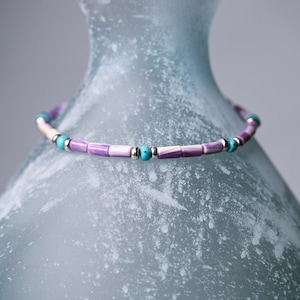 May include: A purple, white, and turquoise beaded necklace with silver accents rests on a textured, frosted glass vase.