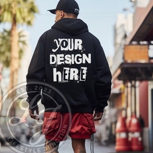 May include: Black hooded sweatshirt with the text "YOUR DESIGN HERE" printed on the back. The sweatshirt is worn by a person wearing red shorts and a black baseball cap. The text "EST. 2023" is printed on the left sleeve of the sweatshirt.