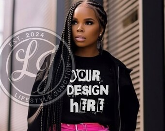 Op de afbeelding: Een zwart T-shirt met de tekst "YOUR DESIGN HERE" in wit. Het shirt wordt gedragen door een persoon met lange zwarte vlechten en een roze broek. De persoon staat voor een gebouw met een wit-zwarte gestreepte muur.