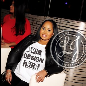 May include: A woman wearing a white t-shirt with the text "YOUR DESIGN HERE" in black lettering. She is wearing a black leather jacket and has long black hair. The woman is sitting in a chair with a white seat. The background is a dark room with a brick wall.