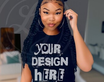 May include: Black t-shirt with the text "YOUR DESIGN HERE" in a distressed white font. The model is wearing large silver hoop earrings and has long braided hair styled in space buns.