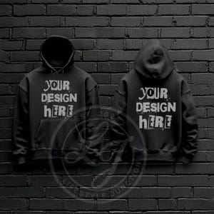 以下が含まれることがあります： 黒いフード付きスウェットシャツ2枚。前面に白い文字で「YOUR DESIGN HERE」と印刷されています。スウェットシャツはレンガの壁にかけられています。