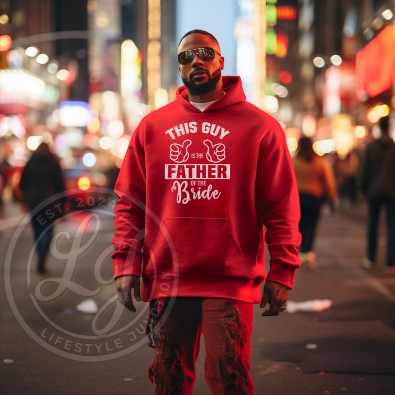 May include: A red hooded sweatshirt with the text "THIS GUY is the FATHER of the Bride" in white. The sweatshirt has a front pocket and a drawstring hood. The image is set against a blurred city background.