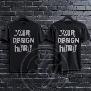 May include: Two black t-shirts with short sleeves hanging on hangers against a black brick wall. The front of each shirt says "YOUR DESIGN HERE" in a distressed white font.
