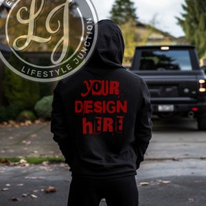May include: Black hoodie with the text "YOUR DESIGN HERE" in red. The hoodie has a hood and is worn by a person. The Lifestyle Junction logo is visible in the upper left corner. A black truck is in the background.