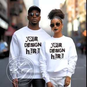 May include: Two white sweatshirts with the text "Your design here" printed in black. The sweatshirts are worn by two people standing in front of a city street background. The sweatshirt on the left has the text "Est. 2023" and a logo below the text.