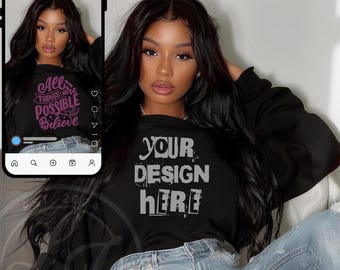 Op de afbeelding: Een zwart sweatshirt met de tekst "YOUR DESIGN HERE" in een wit, verweerd lettertype. Het sweatshirt wordt gedragen door een persoon met lang zwart haar en wordt gecombineerd met een blauwe jeans.